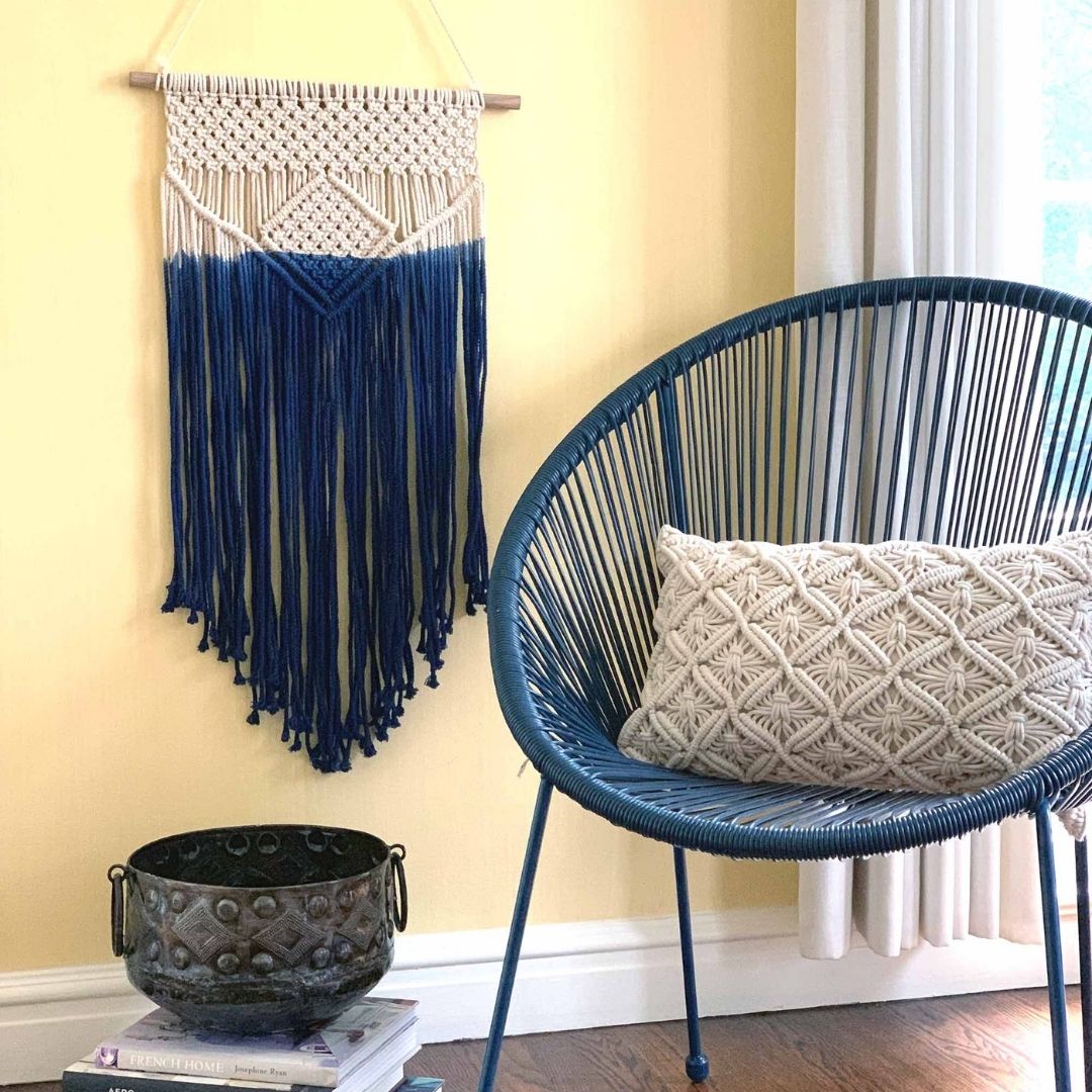 handmade macrame wall hanging in natural and dark blue along side a chair with matching natural colored macrame long pillow cover. Handmade and fair trade imported from New Delhi, India.